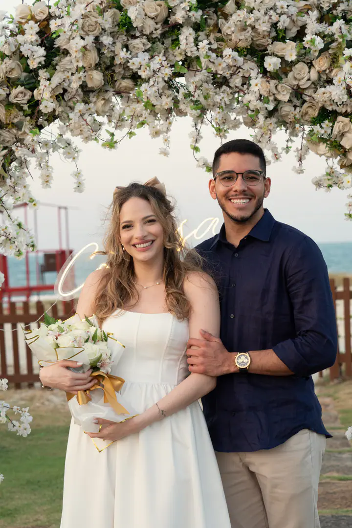 Juliana e Davi sorriem para a câmera sob o arco de flores; ela segura um buquê