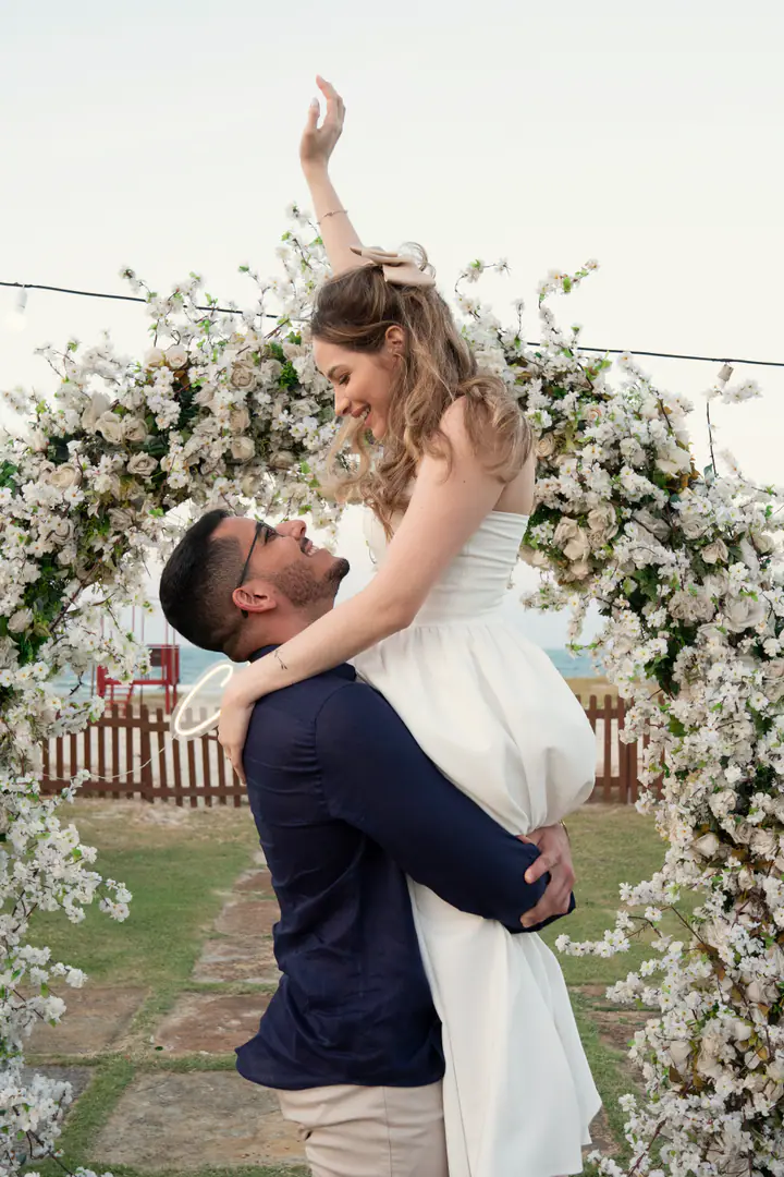 Davi levanta Juliana no colo diante do arco de flores; ela ergue o braço, feliz